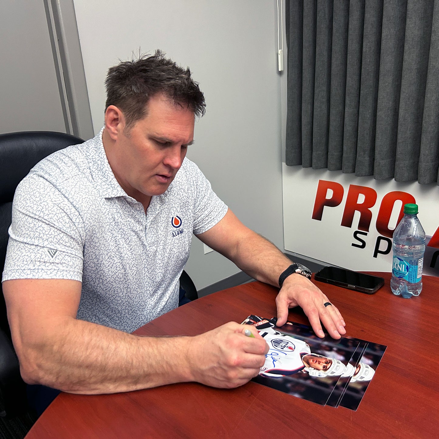 Louie DeBrusk in a white patterned polo shirt sits at a wooden desk, autographing a printed photo. A Pro Sports sign is visible on the wall behind him, along with a water bottle and a black chair.