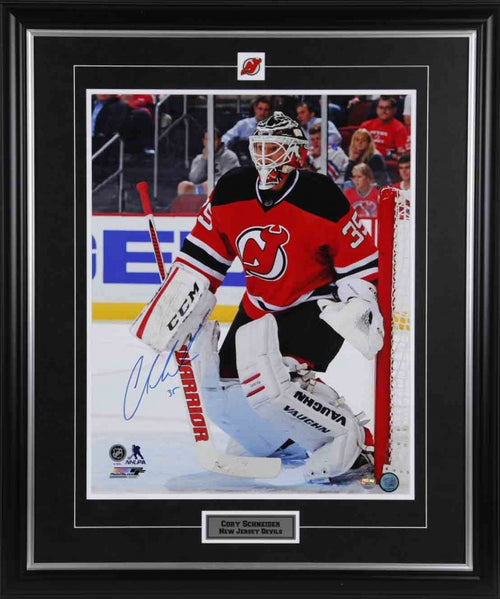 Corey Schneider in the net during a New Jersey Devels hockey game. He is wearing red Devils jersey with white goalie pads. The photo is signed in the bottom left corner using blue ink. Photo is shown framed, with black framing, black mat, inset New Jersey Devils team pin and description plate. 