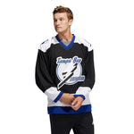 Model wearing the  1992 Tampa Bay Lightning jersey featuring black, white and blue colour blocking and large embroidered logo with circle and lightning bolt. Model is adjusting his sleeve slightly and looking right while walking. He has short wavy blonde hair. 