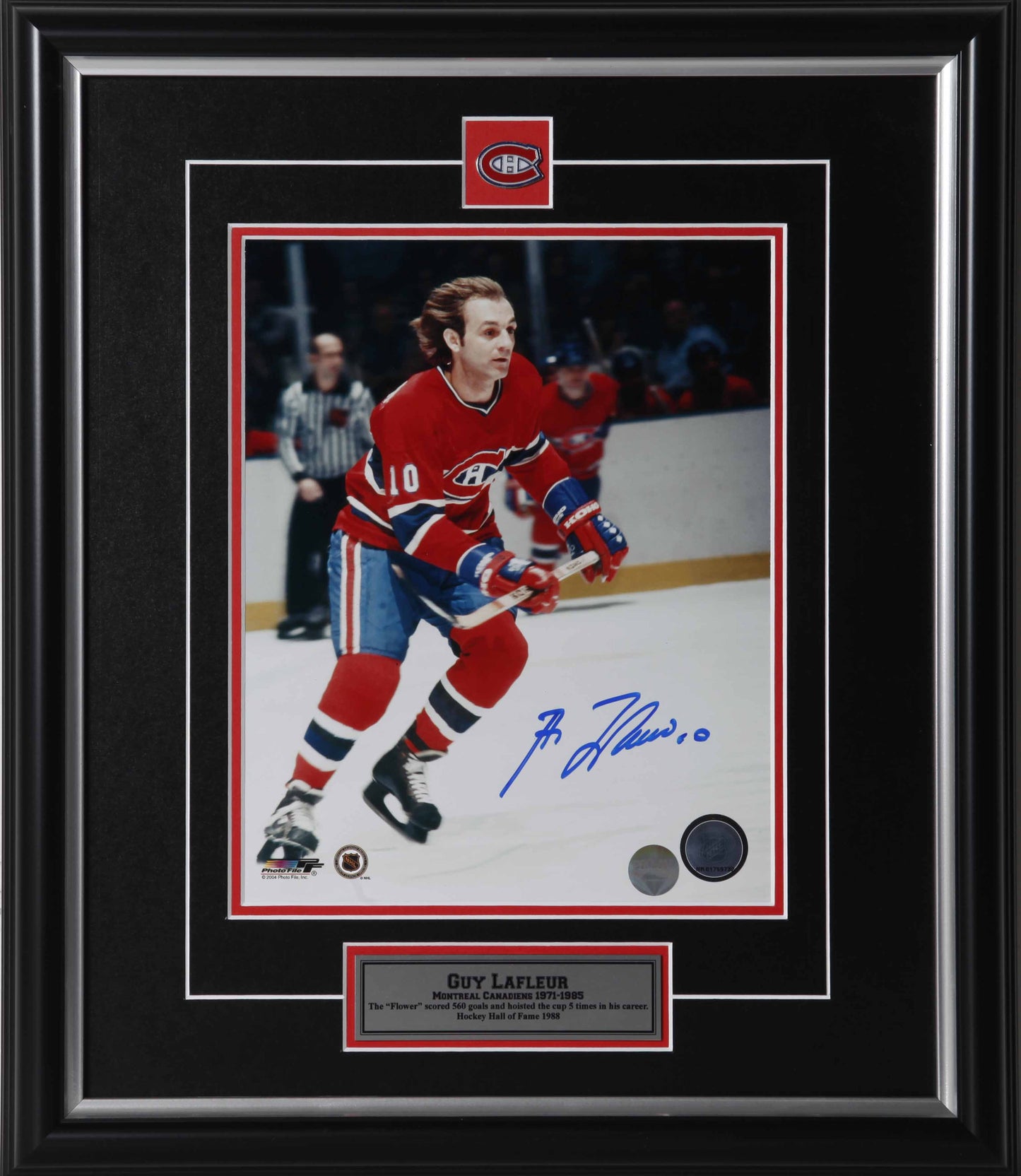 Guy Lafleur skating during a Montreal Canadiens hockey game. He is carrying his hockey stick and wearing red and blue jersey with no helmet. Photo is signed by Lafleur in blue ink. Photo is shown framed, with black framing, black mat with red accent, with inset Montreal Canadiens team pin and description plate