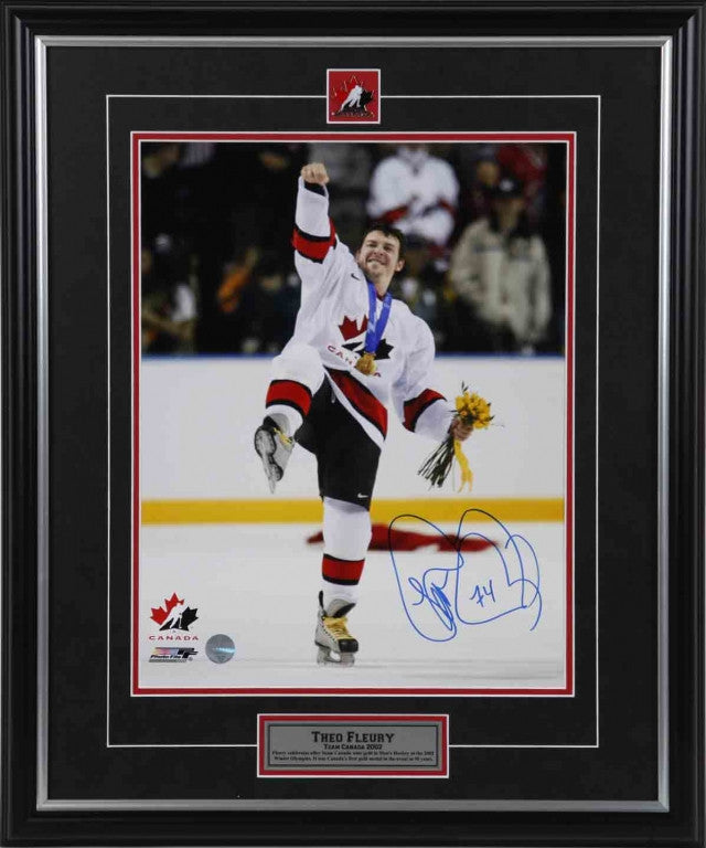 Theo Fleury celebrating Team Canada win with right fist pumped in air and leg up, holding yellow flowers in left hand and wearing medal. Signed in blue in in the bottom right corner. Shown framed, with black frame, black mat with red accents, with Team Canada pin and description plate inset