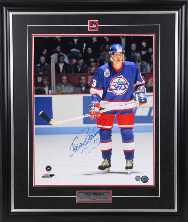 Teemu Selanne during a Winnipeg Jets NHL hockey game, he is wearing blue Jets jersey and is stationery on the ice, holding his stick and watching down ice. Photo is signed by Selanne in blue ink in the bottom left. Image is shown framed, with black frame, black mat with red accent, and inset team pin and description plate.