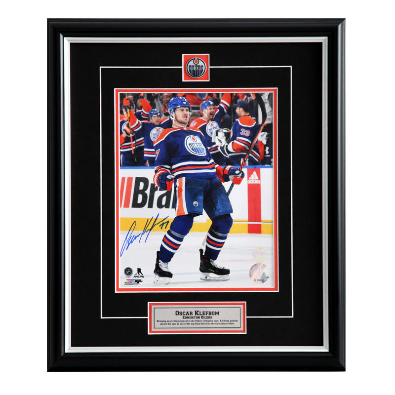 Photo of Oscar Klefbom celebrating during an Edmonton Oilers NHL hockey game. He is wearing the royal blue jersey. Photo is signed in the bottom left corner with blue ink. Photo is shown framed with black framing, black mat with orange accents, and inset team pin and description plate.