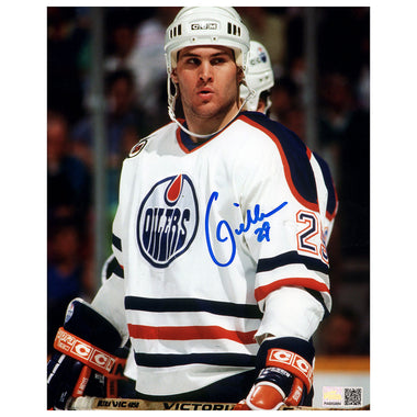 An autographed 8x10 photograph of hockey player Louie DeBrusk in an Edmonton Oilers white jersey, signed in blue ink across the chest. The photo includes a DeBrusk helmet and hockey gear, with a small authentication sticker in the bottom right corner.
