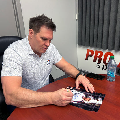 Louie DeBrusk in a white patterned polo shirt sits at a wooden desk, autographing a printed photo. A Pro Sports sign is visible on the wall behind him, along with a water bottle and a black chair.