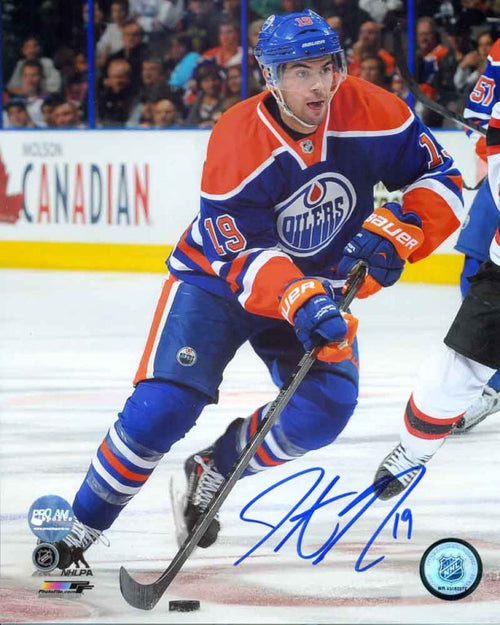 Photo of Edmonton Oiler Justin Schultz skating with the puck during an Oilers NHL game, wearing royal blue jersey. Photo is signed with blue ink in the lower right corner. 