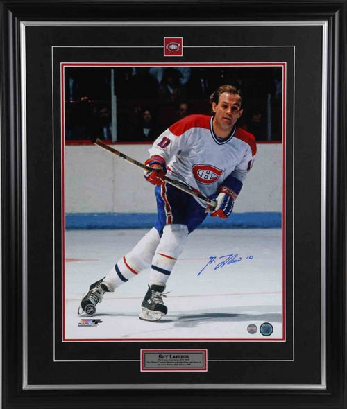 Guy Lafleur skating during a Montreal Canadiens hockey game. He is wearing white Canadiens jersey and no helmet. The photo is signed in the bottom right corner in blue ink. Image is shown framed, with black framing, black mat with red accent, and inset Calgary Flames team pin and description bar. 