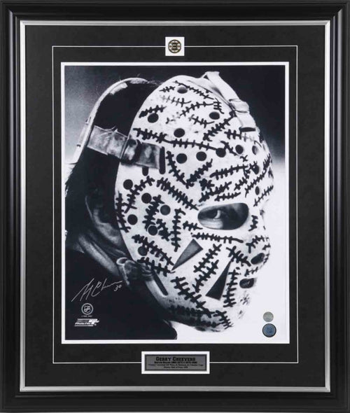 Portrait photo of Gerry Cheevers of the Boston Bruins wearing his famous goalie masks with drawn on "stitches". The photo is in black and white; signed by Cheevers with silver ink in the bottom left corner.  Photo is shown framed, with black frame and black mat, inset Boston Bruins team pin and the top and description bar in the bottom  
