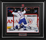 Brendan Gallagher in front of net during a Montreal Canadiens NHL hockey game. He is facing away from the camera, his legs wide and arms in air. He is wearing white team jersey. Image is signed by Gallagher in blue with on the right side of the photo.  Photo is shown framed, with black frame, black mat with red accent, and inset Montreal Canadiens team pin and description plate. 