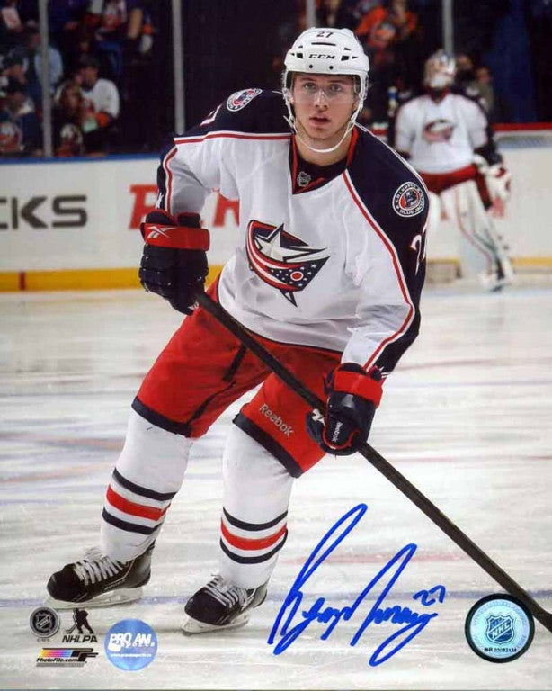 Ryan Murray of the Columbus Blue Jackets skating with hockey stick during an NHL hockey game. He is wearing white jersey with red shorts. He is looking directly at the camera. Photo is signed by Murray in blue ink in the bottom right corner. 