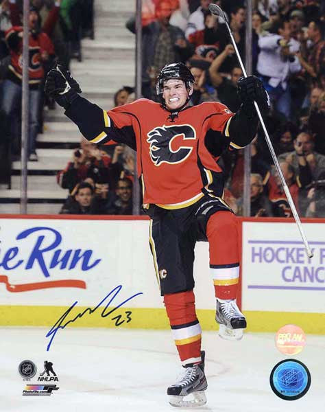 Sean Monahan celebrating during a Calgary Flames NHL hockey game. He is wearing red jersey with black shorts and helmet. Photo is signed by Monahan in the bottom left corner
