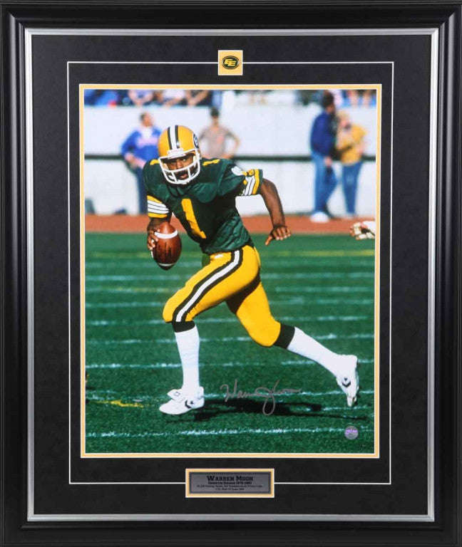 Warren Moon Edmonton Elks / Edmonton Football Team shown running with the ball, wearing green and gold uniform. Image is signed by Moon with silver in at the bottom of the photo. Photo is shown framed, with black frame, black mat with golden yellow accent, and inset Edmonton Football Team pin at top and  description plate at bottom. 