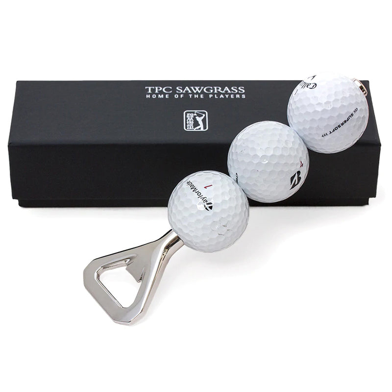 Bottle opener made from three game used golf balls collected from TPC Sawgrass, opener is photographed leaning against black gift box with silver "TPC Sawgrass, Home of the Players" and logo. 