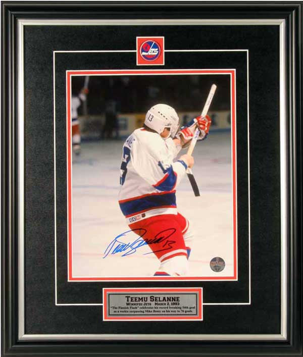 Teemu Selanne celebrating his Rookie Goal Record during a Winnipeg Jets NHL game. He is facing away from the camera and is wearing the Jets' white jersey. The photo is signed by Selanne in blue ink in the bottom centre.  Image is shown framed, with black frame, black mat with red accent, and inset Winnipeg Jets team pin and descriptive plate. 