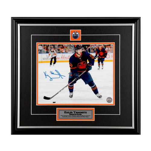 Photo of Edmonton Oiler Kailer Yamamoto skating with puck during an NHL hockey game; he is wearing navy jersey. Photo is signed in blue in the lower left. Photo is shown framed with black framing and mat with orange accents, inset team pin and description plate. 