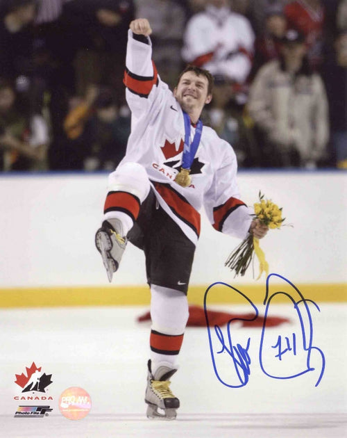 Theo Fleury celebrating Team Canada win with right fist pumped in air and leg up, holding yellow flowers in left hand and wearing medal. Signed in blue in in the bottom right corner. 