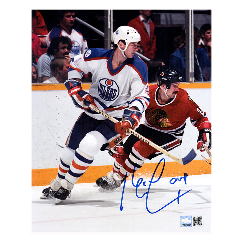 Action shot of Kevin Lowe skating during an Edmonton Oilers NHL game against Chicago. Lowe is looking over his left shoulder and opponent is skating closely behind him. Lowe is wearing white Oilers jersey. Image is signed by Lowe in blue ink in the bottom right