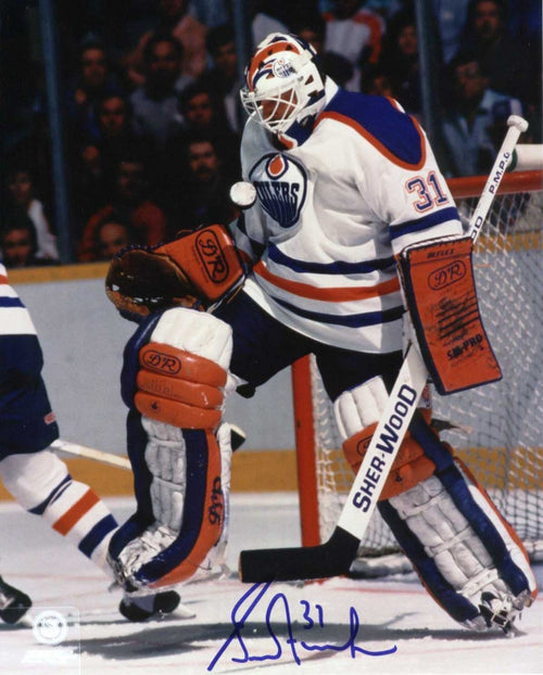 Edmonton Oilers goalie Grant Fuhr in front of the net during an Oilers game. He is wearing a white team jersey and white, blue and orange goalie pads. Photo is signed by Fuhr in blue ink at the bottom centre of the image. 