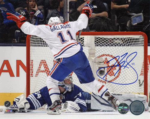 Brendan Gallagher in front of net during a Montreal Canadiens NHL hockey game. He is facing away from the camera, his legs wide and arms in air. He is wearing white team jersey. Image is signed by Gallagher in blue with on the right side of the photo. 