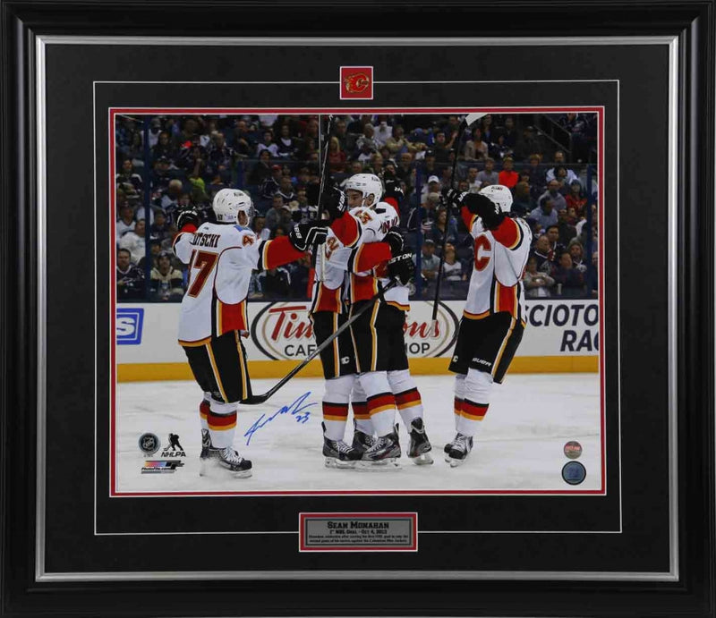 Sean Monahan celebrating with his Calgary Flames teammates on ice after his first NHL goal. Image is signed in blue ink by Monahan. Photo is showed framed with black frame, black mat with red accent, and inset Calgary Flames team pick and description bar. 