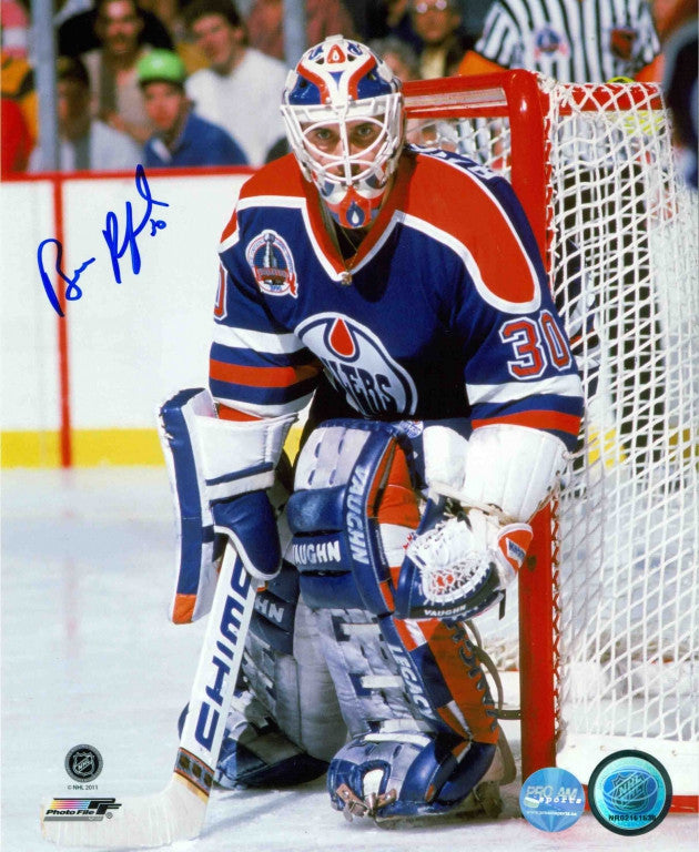 Bill Ranford guarding net during an Edmonton Oilers hockey game. Ranford is wearing royal blue Oilers jersey and blue goalie pads. He is staring directly into the camera. Photo is signed on the left side near his shoulder. 