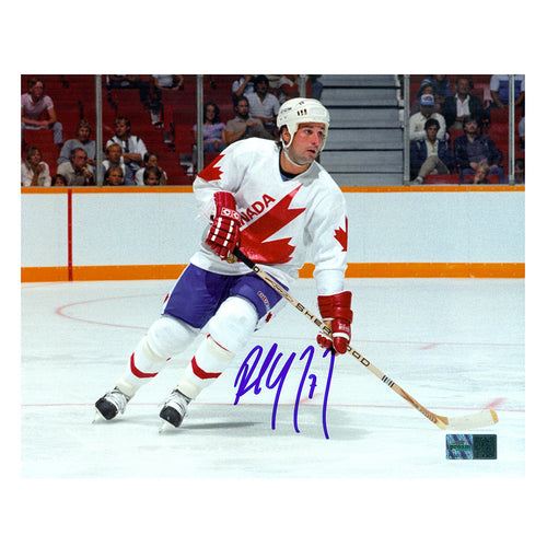 signed photo of Paul Coffey Team Canada Skating during a game wearing white Team Canada jersey. He is looking to the left down the rink. Photo is signed in the lower center in blue ink. 