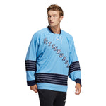 Model wearing the adidas vintage team classics 1967 Pittsburgh Penguins jersey, featuring blue and white striping on sleeves and lower hem. Model has short wavy blonde hair and he is looking to the right while walking