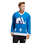 Model wearing the adidas vintage team classics 1979 Quebec Nordiques jersey, featuring the igloo with leaning hockey stick logo and fleur de lis. the model is looking left and walking; he has short blonde wavy hair. 