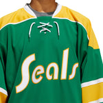 Detail front view of the adidas vintage team classics 1970 California Golden Seals jersey featuring yellow and white text logo. Jersey is primarily green with bands of yellow and white. Collar has lace up detail.