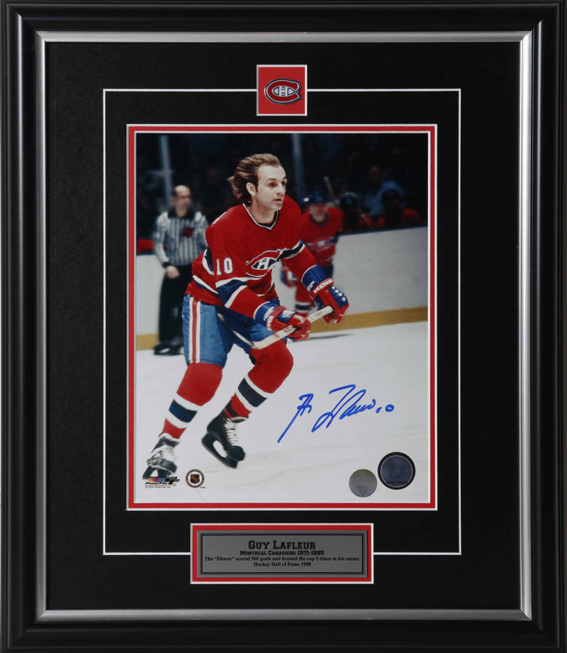 Guy Lafleur skating during a Montreal Canadiens hockey game. He is carrying his hockey stick and wearing red and blue jersey with no helmet. Photo is signed by Lafleur in blue ink.  Photo is shown framed, with black framing, black mat with red accent, with inset Montreal Canadiens team pin and description plate