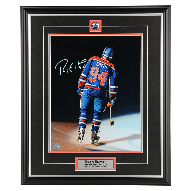 Photo of Edmonton Oilers Ryan Smyth skating away towards dark background, wearing royal blue jersey, during his last game with the Oilers before retirement. Photo is signed in light blue ink in the upper left. Photo is shown framed, with black framing and mat with orange accents and inset team pin and description plate. 