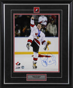 Theo Fleury celebrating Team Canada win with right fist pumped in air and leg up, holding yellow flowers in left hand and wearing medal. Signed in blue in in the bottom right corner. Shown framed, with black frame, black mat with red accents, with Team Canada pin and description plate inset