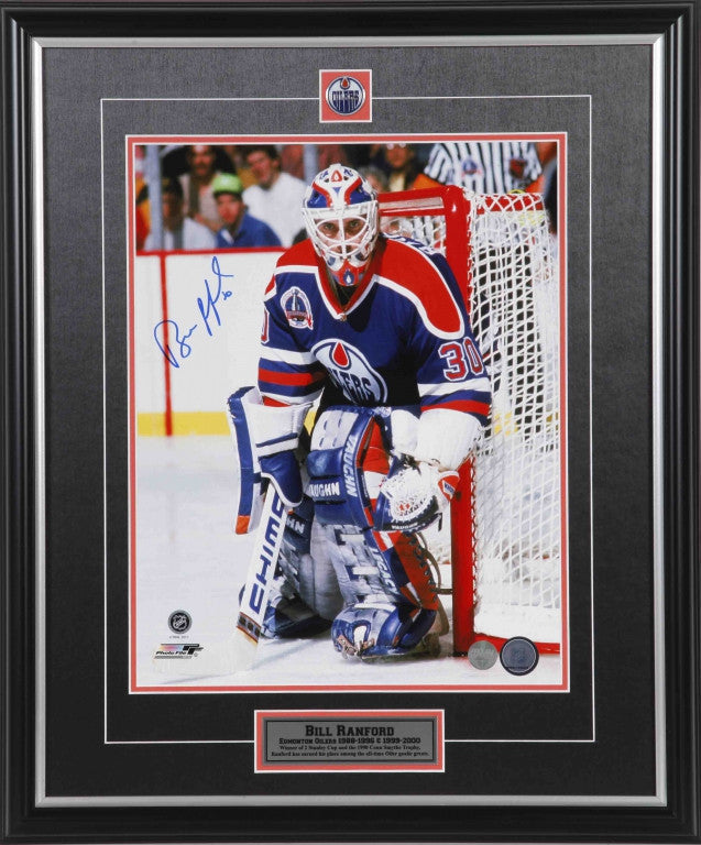 Bill Ranford guarding net during an Edmonton Oilers hockey game. Ranford is wearing royal blue Oilers jersey and blue goalie pads. He is staring directly into the camera. Photo is signed in blue ink on the left side near his shoulder. Photo is shown framed with black framing, black mat with orange accent, and inset Edmonton Oilers team pin and description plate. 