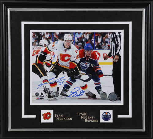 Photo of Ryan Nugent-Hopkins (Edmonton Oilers) and Sean Monahan (Calgary Flames) during an NHL hockey game. Photo is dual signed by Sean Monahan and Ryan Nugent-Hopkins, signatures are done in blue ink. Photo is shown framed in black framing and mat with white accents. Team pins are inset beside players names. 