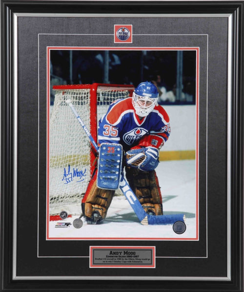 Andy Moog photographed guarding the net during an Edmonton Oilers NHL hockey game. Moog is wearing royal blue Oilers jersey with blue and brown pads. Photo is signed on the left side near the bottom in blue ink. Photo is showed framed, with black framing, black mat with orange accents, and inset Edmonton Oilers team pin and description plate. 