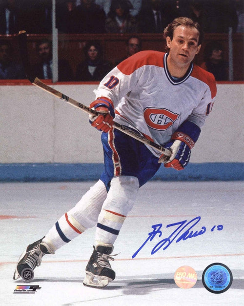 Guy Lafleur skating during a Montreal Canadiens hockey game. He is wearing white Canadiens jersey and no helmet. The photo is signed in the bottom right corner in blue ink. 