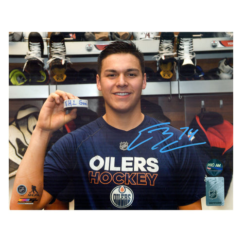 Photo of Ethan Bear in locker room holding hockey puck with visible text "NHL Goal" written on it. Photo is signed in light blue in in the lower right