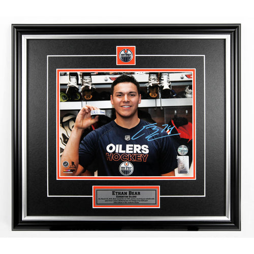 Photo of Ethan Bear in locker room holding hockey puck with visible text "NHL Goal" written on it. Photo is signed in light blue in in the lower right. Photo is shown framed, with black frame and mat with orange accents and inset team pin and description bar. 